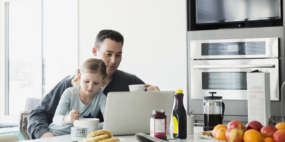 Un homme travaille devant son ordinateur dans sa cuisine, sa fille sur ses genoux. 