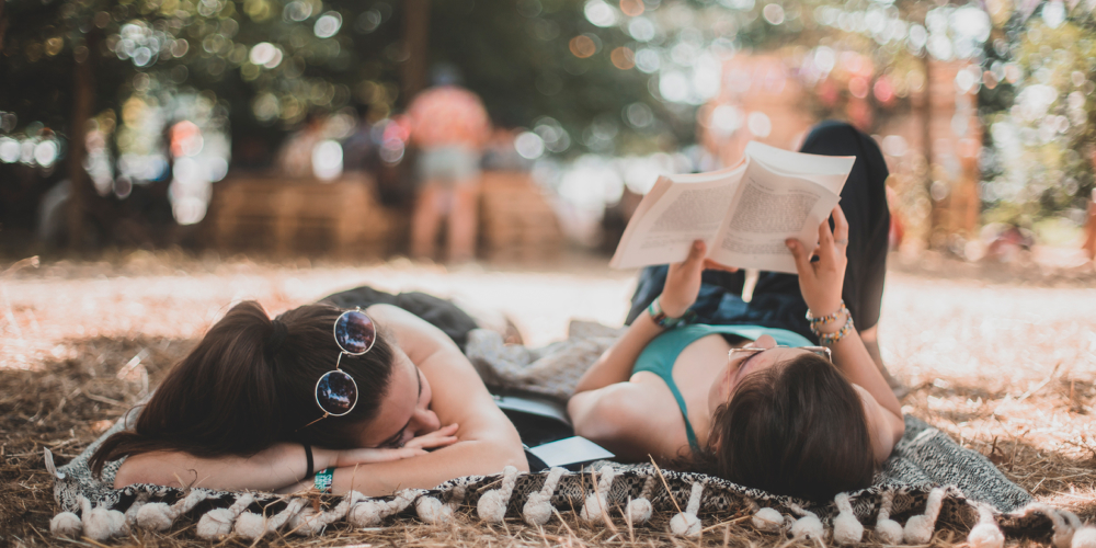 Two young women are reading in a park, lying on a blanket. 

 