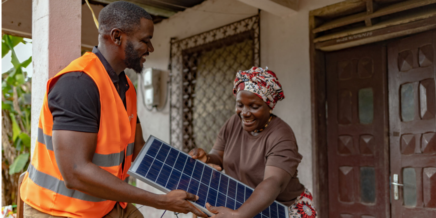 An Orange Energies technician carries a solar panel with an African woman in front of her house. 