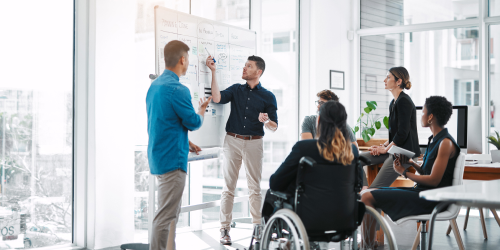 Five people, including one in a wheelchair, listen to a sixth person stationed in front of a whiteboard during a meeting.   