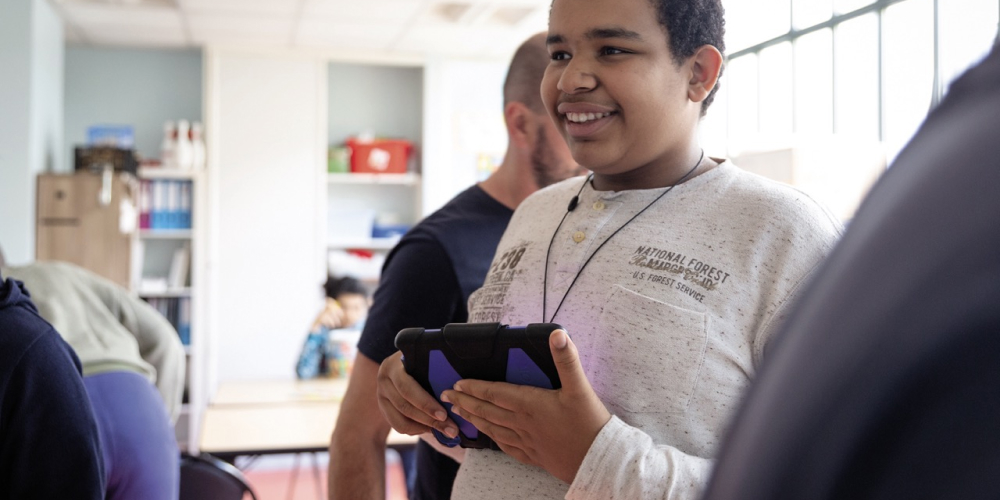 A young autistic boy smiles at people around him, a digital tablet in his hands.