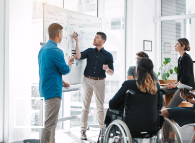 Five people, including one in a wheelchair, listen to a sixth person stationed in front of a whiteboard during a meeting.   
